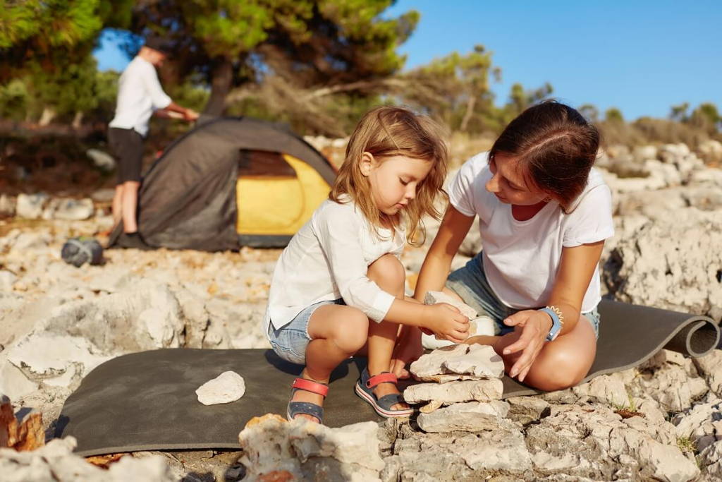 Mother and daughter playing with rocks on the beach while the father is setting up a tent in the background.