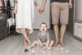 Toddler sitting on the floor while parents stand nearby holding hands, demonstrating calm support