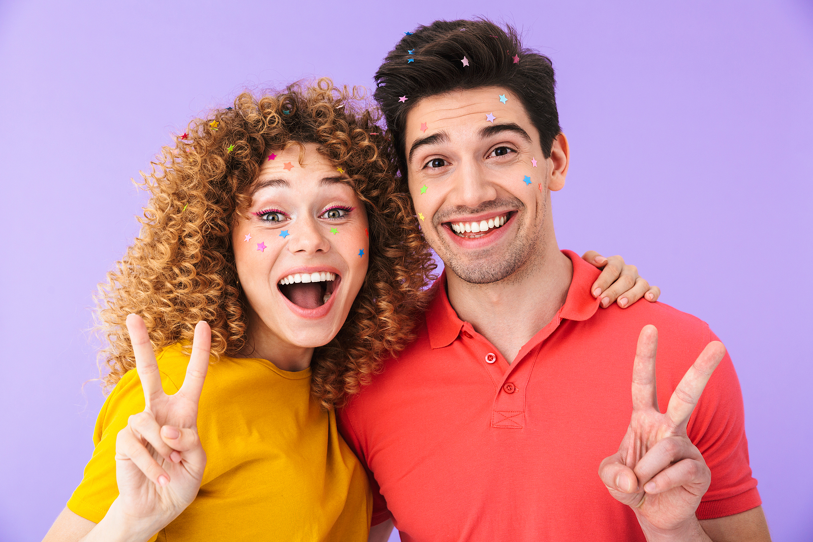 An attractive man and woman smile and hold a peace sign while side hugging.