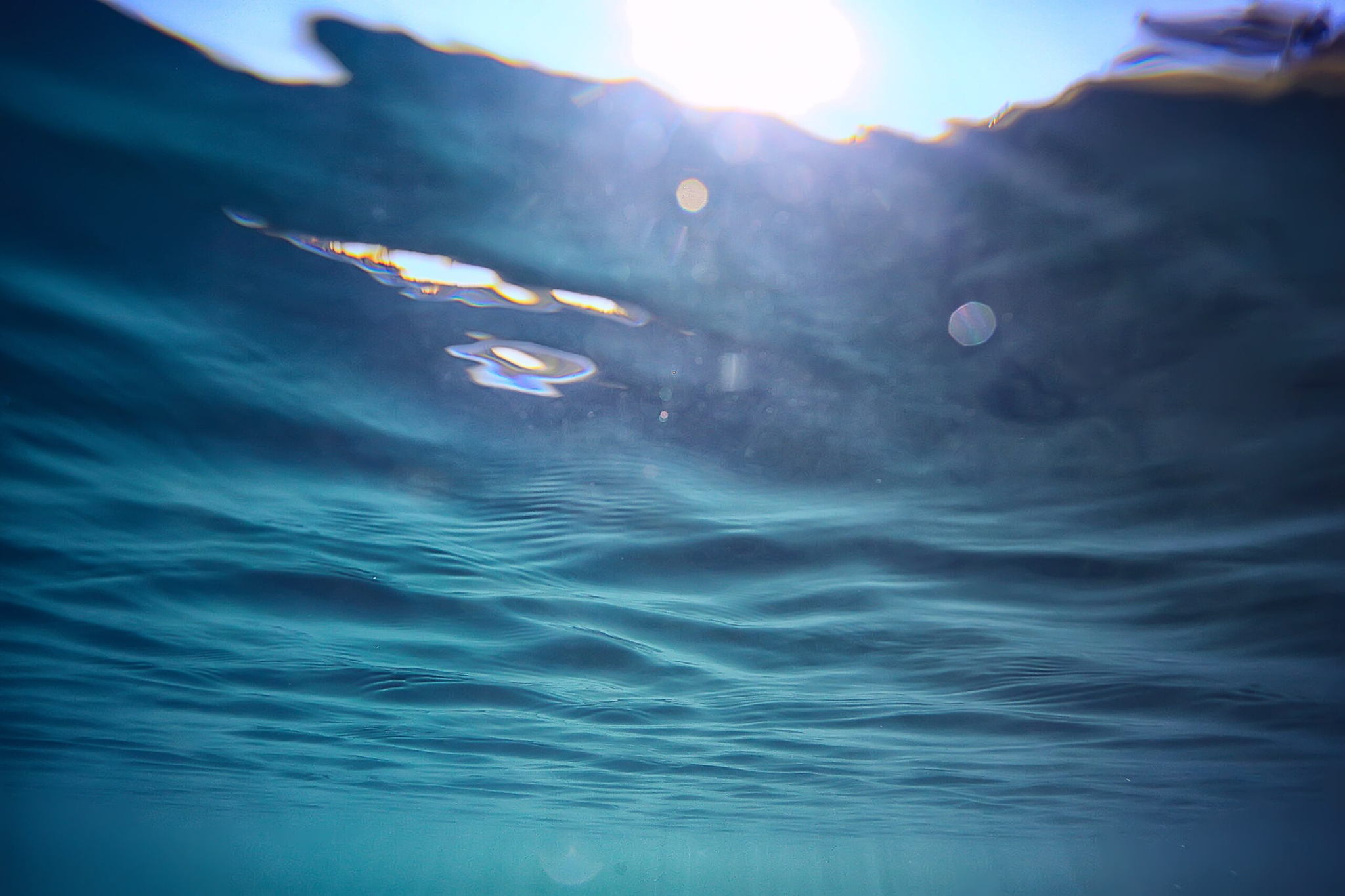 Underwater view looking up at the sunlit ocean surface, with light shining through blue water and gentle waves creating ripples above.