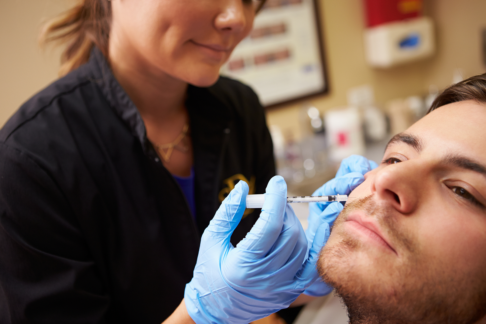 A doctor smiles as she injects a young man with botox underneath his eyes. The young man has a serious expression.