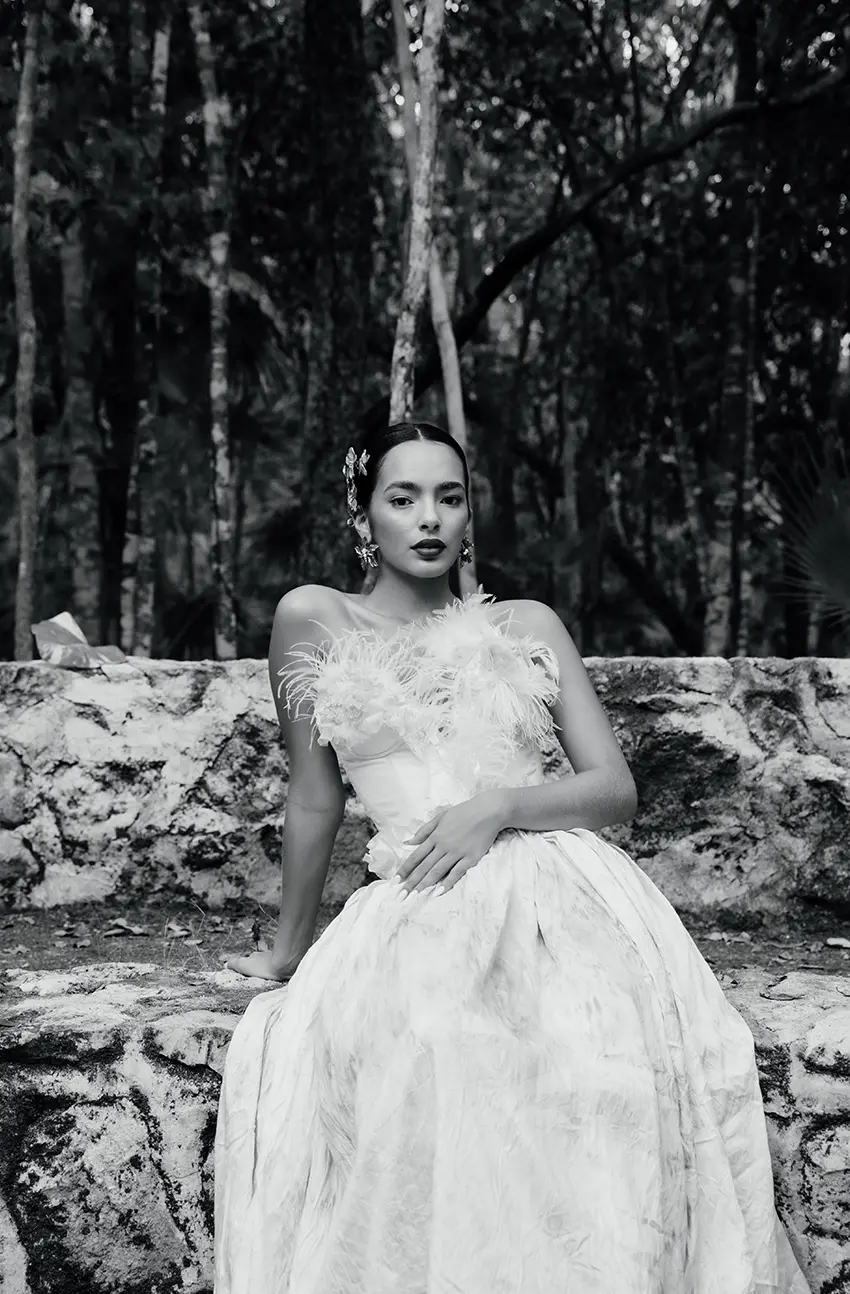 Une mariée dans une élégante robe de mariée blanche ornée de plumes sur les épaules, assise contre un mur de pierre dans un cénote à Tulum.