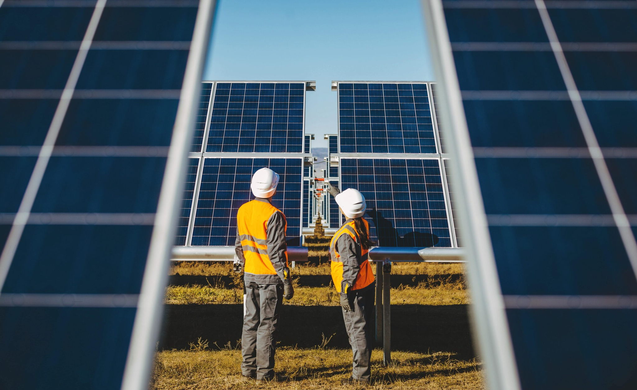 Two workers in orange safety vests and white helmets stand between rows of solar panels, inspecting them. The panels are aligned in a field under a clear blue sky, capturing sunlight for energy.