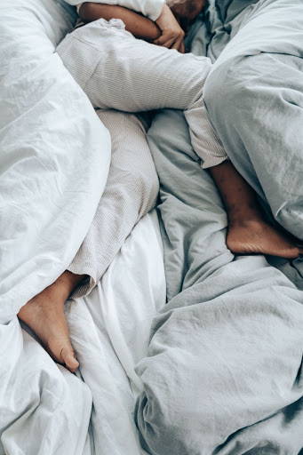 A couple snuggled up in bed, with their legs poking out of the blankets - Photo by Ketut Subiyanto from Pexels