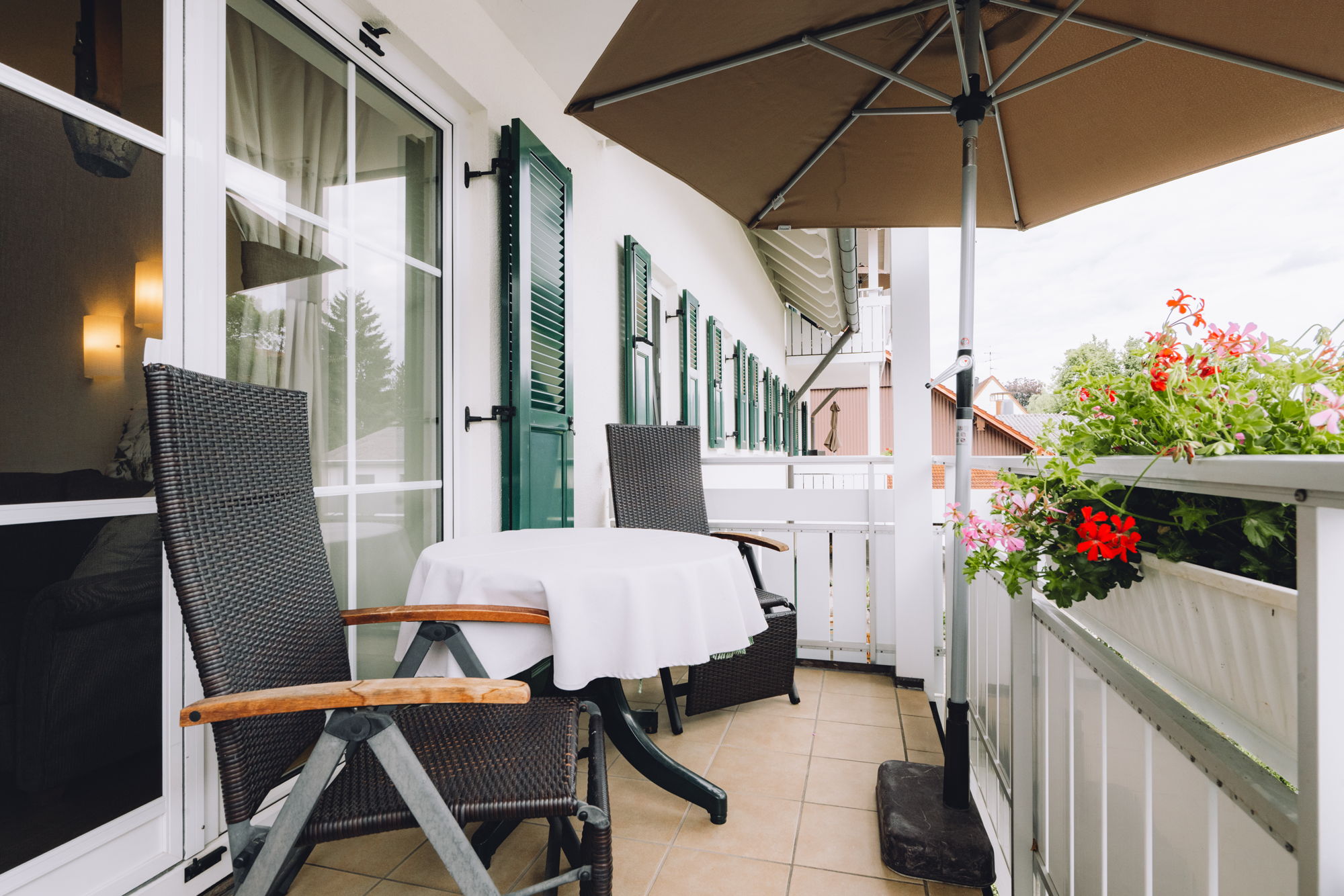 Ein Balkon mit Sonnenschirm, weißem Tisch, verstellbaren Stühlen und Blumenkasten.