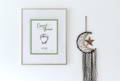 Framed baby footprint keepsake that has newborn baby footprints, baby’s name and birth information hung on wall next to dream catcher decoration, clicking this image leads to the baby footprint collection