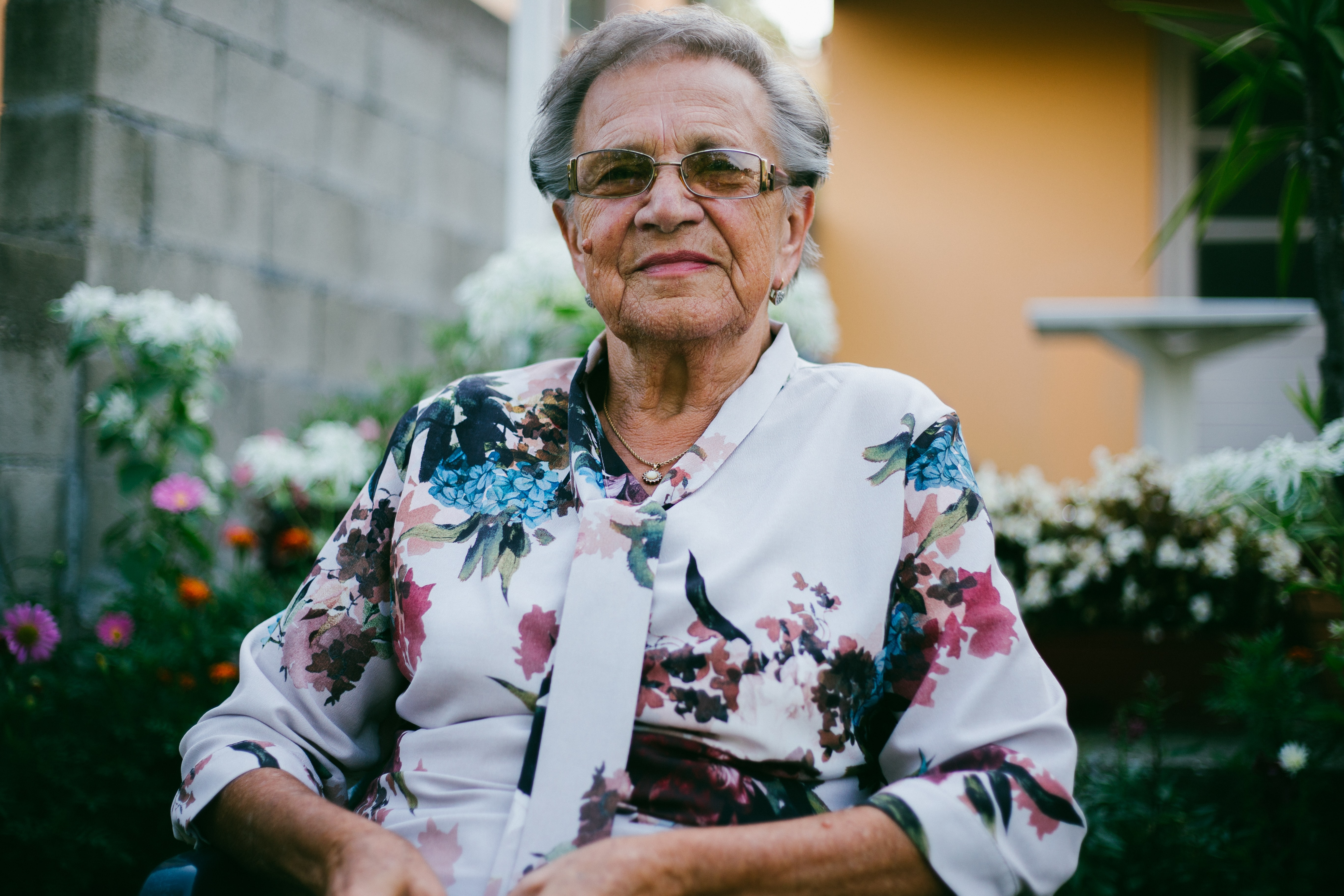 An older woman with flowered shirt sits and smiles in front of her garden.