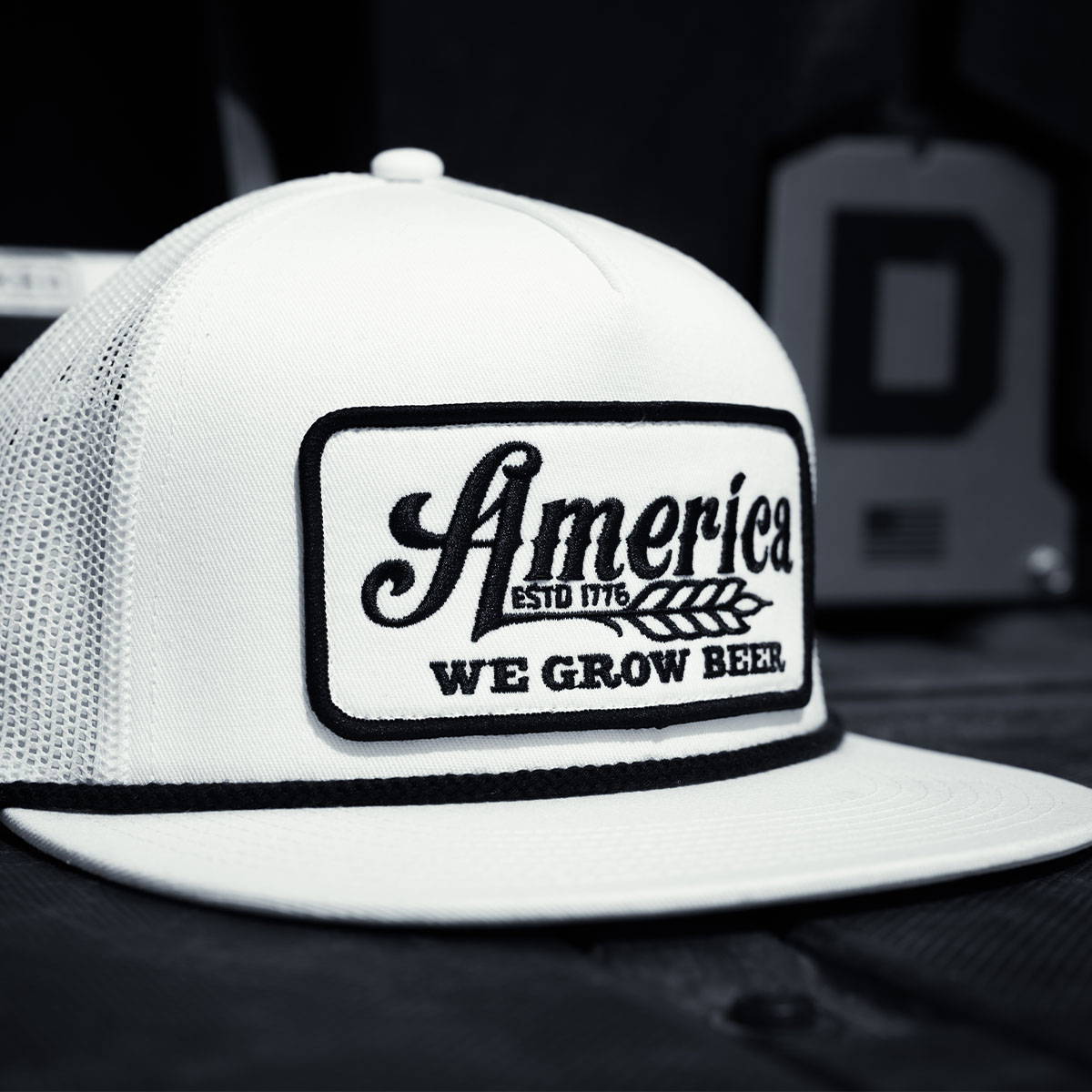 A white flat bill cap with a woven patch that says "America We Grow Beer." Represents the New Arrival Category