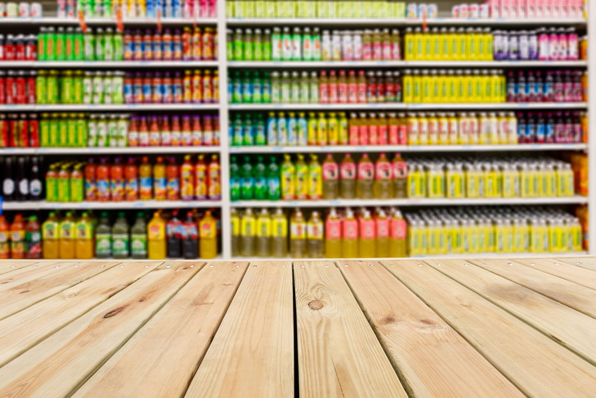 Wooden tabletop in front of blurred supermarket shelves filled with colorful bottles and cans of beverages.