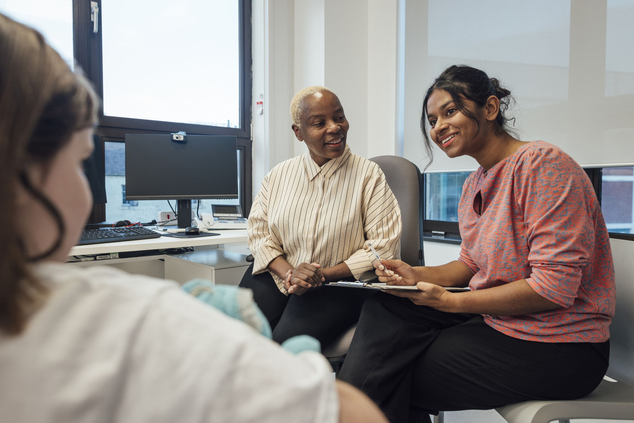 Two women sit in an office, one holding a clipboard and pen, smiling and talking to a person sitting across from them. A computer and office supplies are on the desk by the window in the background.