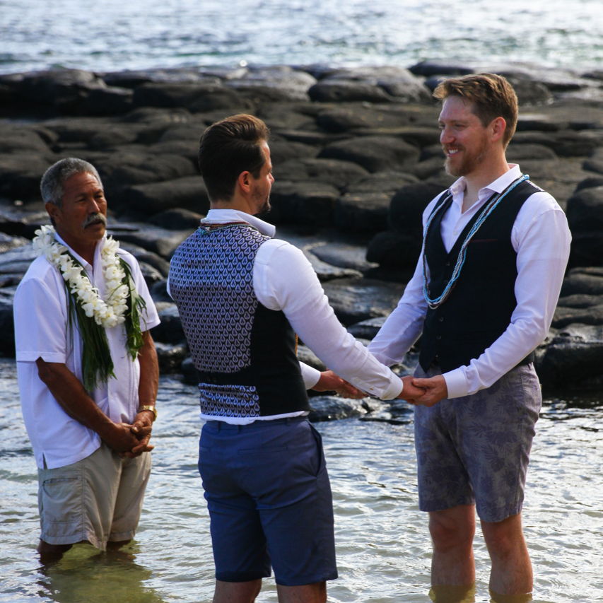 Two men holding hands on the beach, looking into each other's eyes.