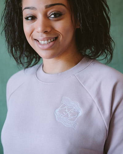 Front of woman wearing dusty pink organic cotton sweatshirt with white embroidered design on the left chest, from women's sustainable clothing brand Birdsong