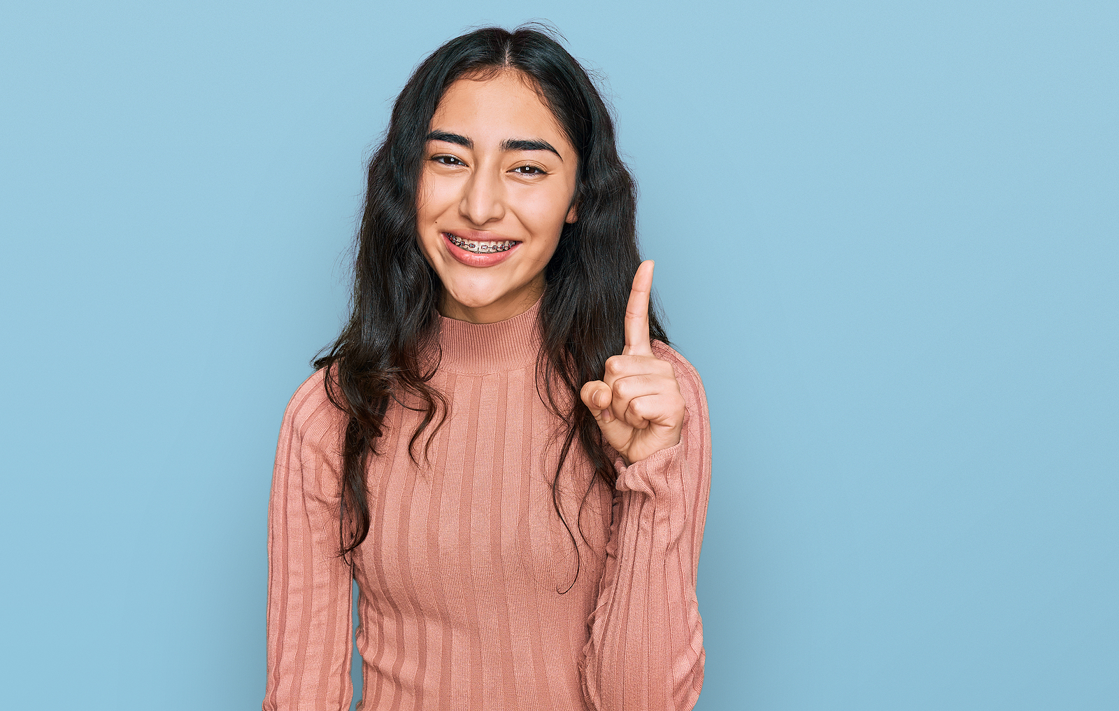 A young woman with braces smiles while holding a finger up against a plain background.