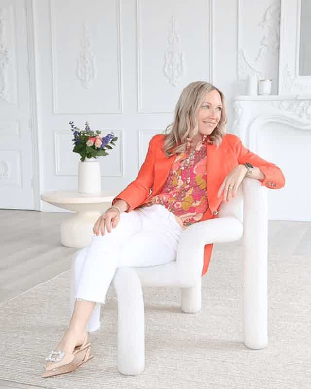 Woman seated in modern white chair, wearing coral blazer and floral blouse, in bright elegant room.