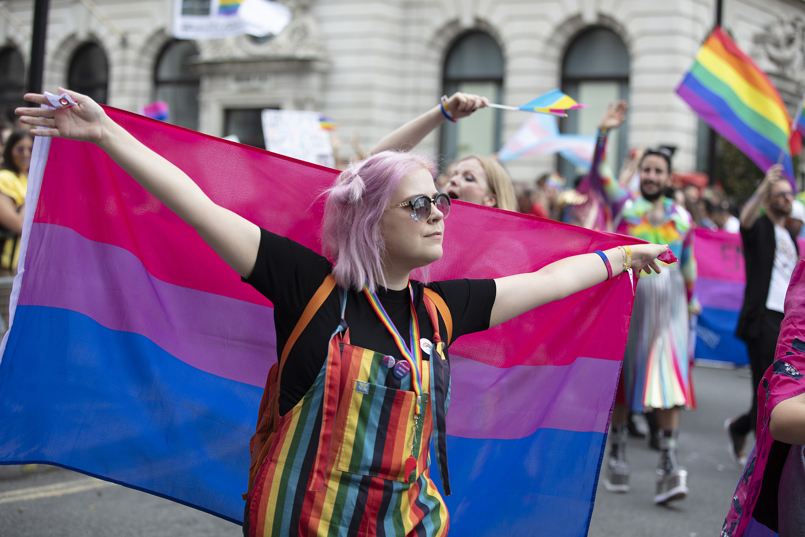 A woman with rainbow overalls holds the pride flag behind her while at pride.