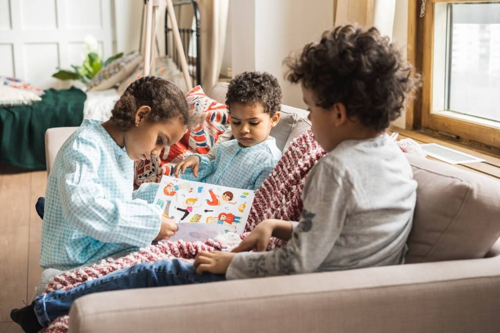 Three young children sitting on a cozy couch by the window, engaged in reading a colorful picture book together on a relaxed indoor day.