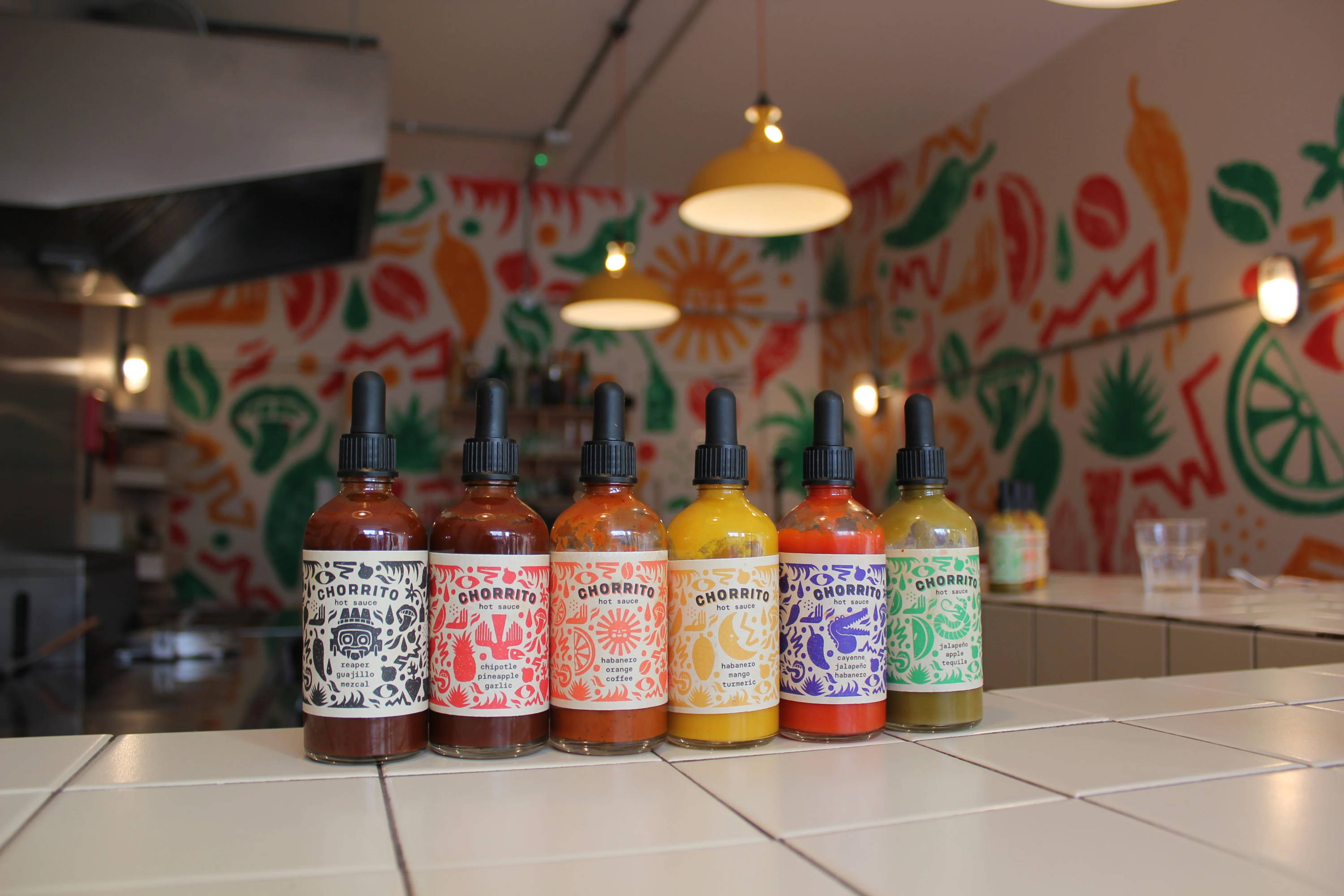 Six colourful bottles of hot sauce sitting atop a white tiled corner bar in a Cantina. In the banground is 2 Coolicon Lampshades hanging above which are out of focus.