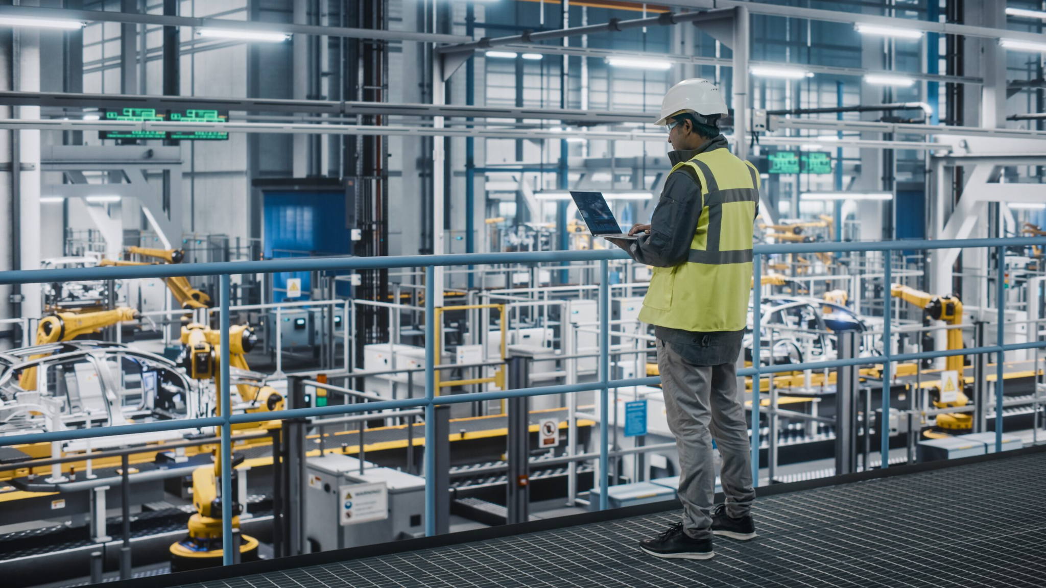 A person wearing a safety vest, hard hat, and mask stands on a balcony in a modern factory, using a laptop and observing robotic machinery on the assembly line below.
