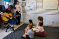 An image of a young boy playing the ukuele with a a satisfied look on his face. He is in front of a large window that and there is a calm, cool day outside. 