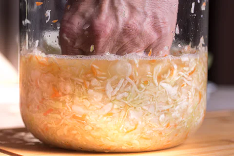 A hand presses finely chopped white cabbage and carrot into a glass jar for fermentation, with liquid and small bubbles visible on the surface.