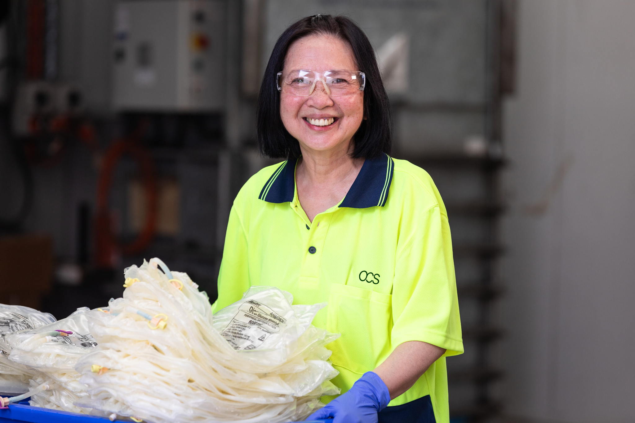 A woman wearing safety glasses, a bright yellow work shirt, and blue gloves smiles while standing next to a pile of packaged medical tubing in an industrial setting.