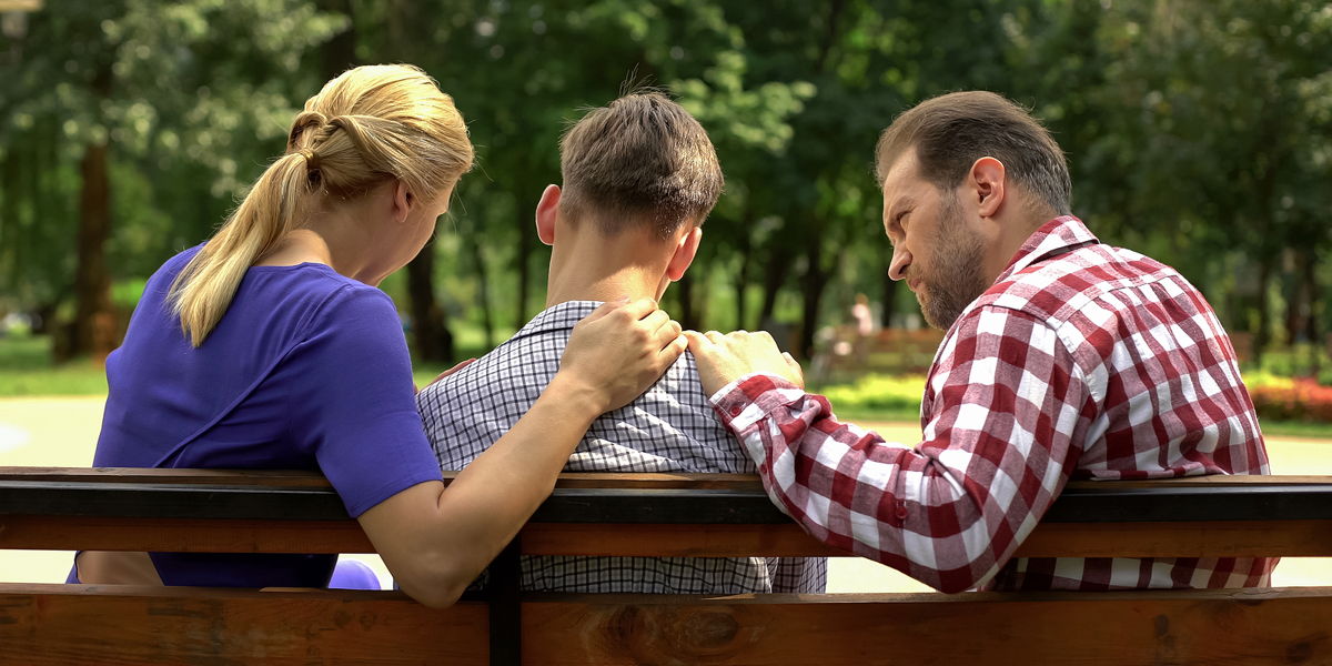 A family of 3 sitting on a bench. The parents have a hand on a young mans shoulders while he tilts his head.