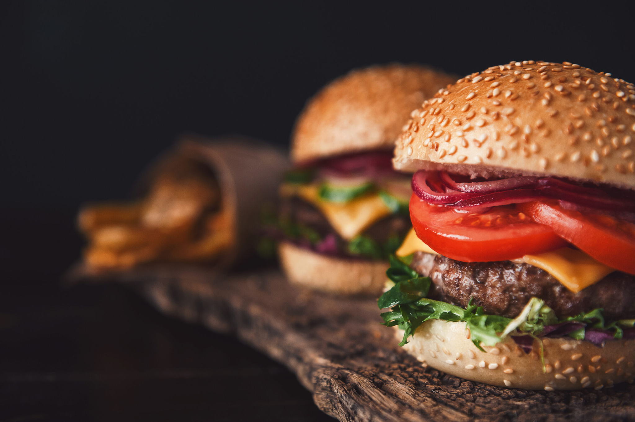 Close-up of two cheeseburgers with lettuce, tomato, red onion, cheese, and sesame seed buns on a rustic wooden board, with a serving of fries in the background.