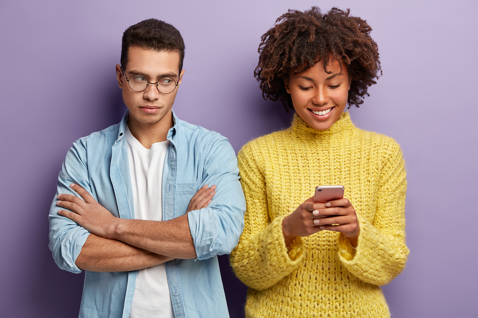 A man with his arms crossed looks over a his friend who is smiling looking down at her phone. He has an annoyed expression.