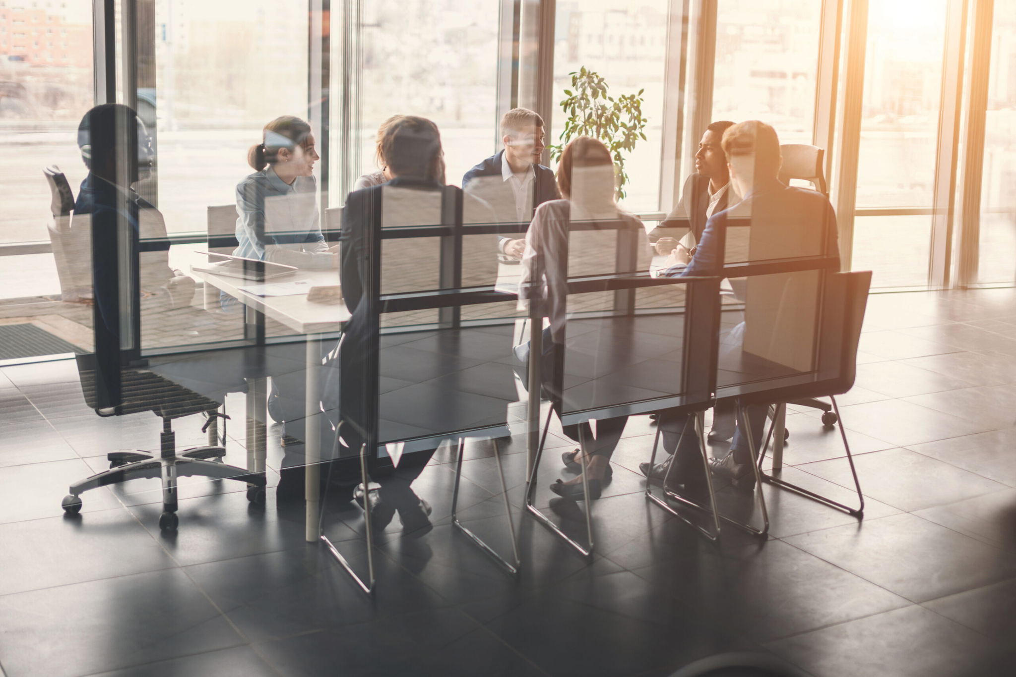 A group of people sit around a table in a modern office conference room with large windows, discussing business. Sunlight streams in, and the scene is reflected on the glass walls.