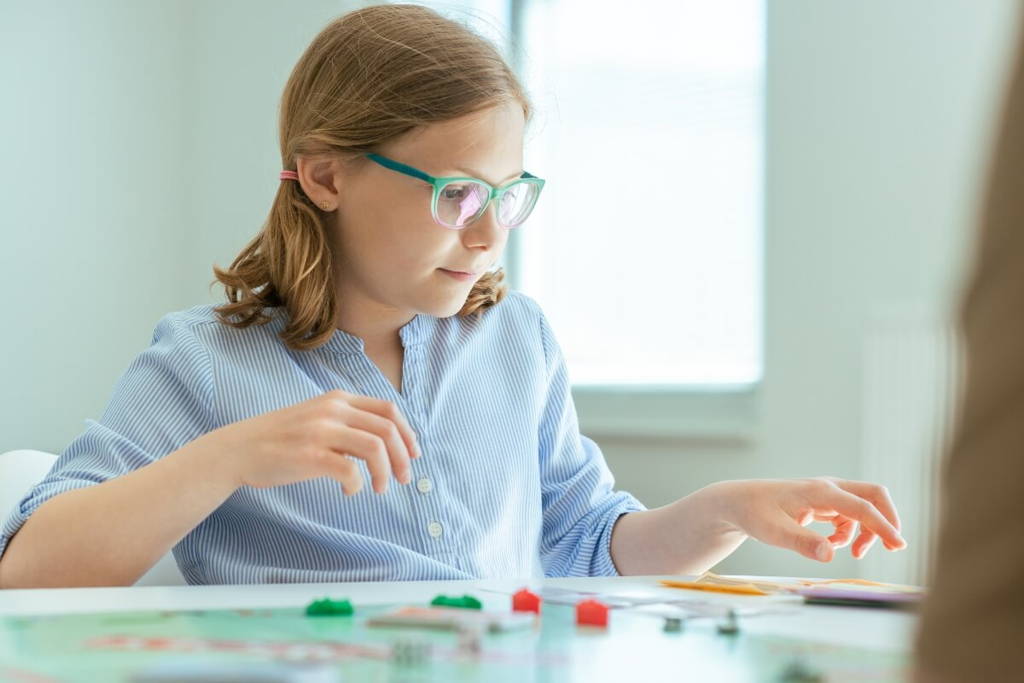 Jeune fille concentrée, portant des lunettes et une chemise à rayures, jouant à un jeu de société à une table dans une pièce bien éclairée.