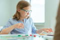 Focused girl wearing glasses and a striped shirt playing a board game at a table in a brightly lit room.
