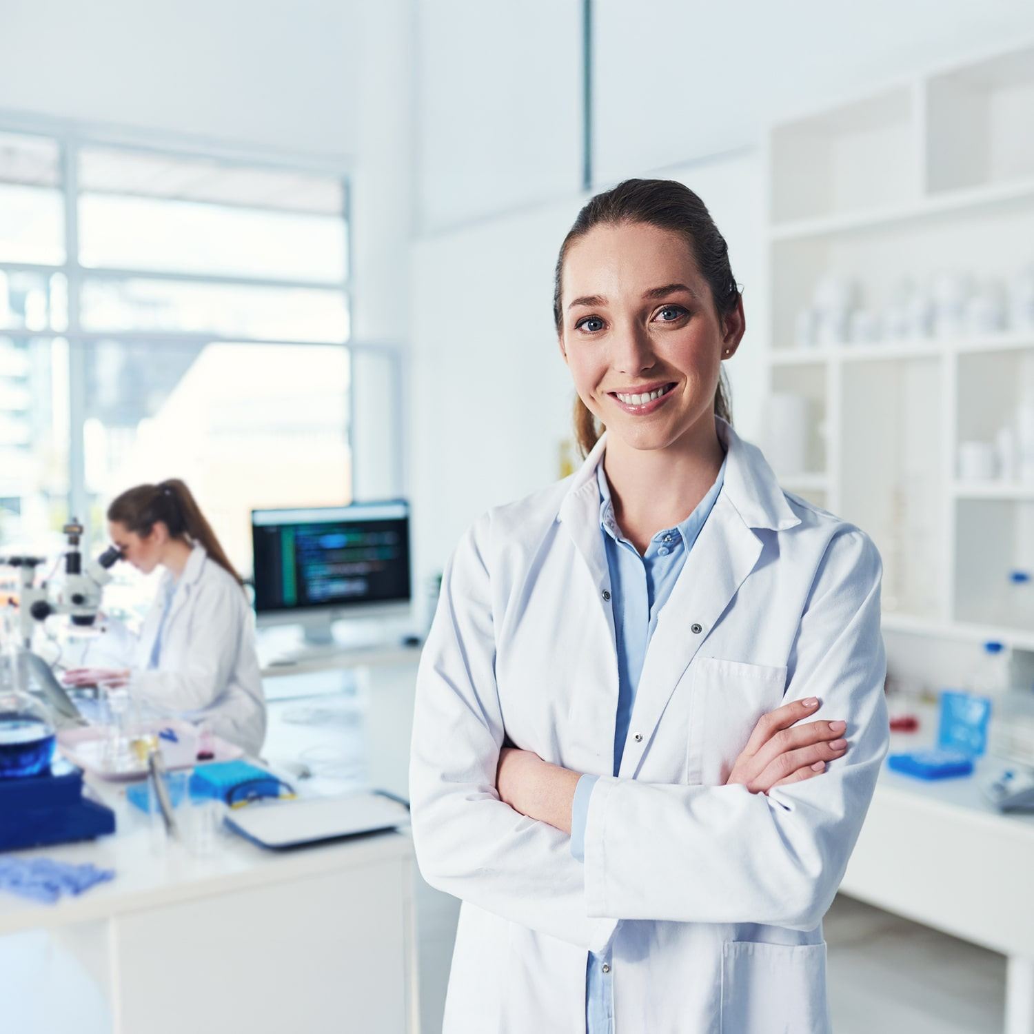 A woman in a white lab coat stands smiling with arms crossed in a bright, modern laboratory, while another person works at a lab bench in the background.