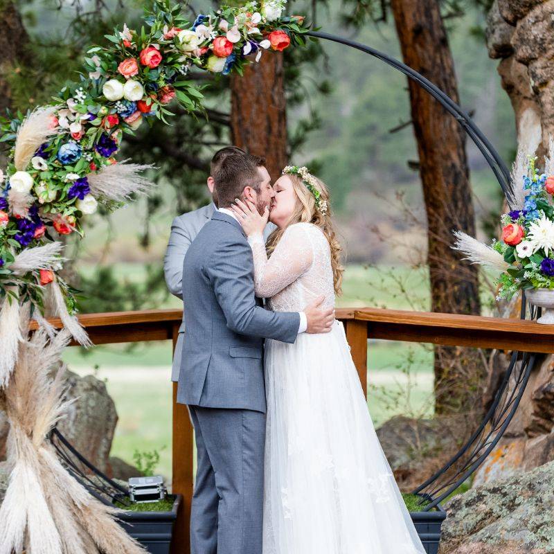 A bride and groom share their first kiss in front of a black circular arch with pampas grass, coral David Austin roses, blue anemones, cream David Austin roses, and greenery