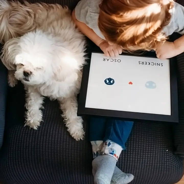 young child holding art with two dog nose prints  sits on couch with small white dog