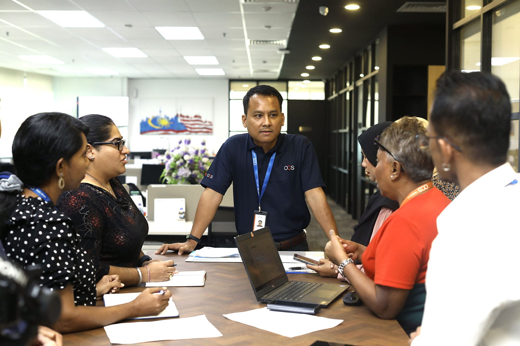 A man stands at the head of a conference table, speaking to five seated colleagues in an office. Laptops, notebooks, and pens are on the table as they have a discussion. The office is modern with glass panel walls.