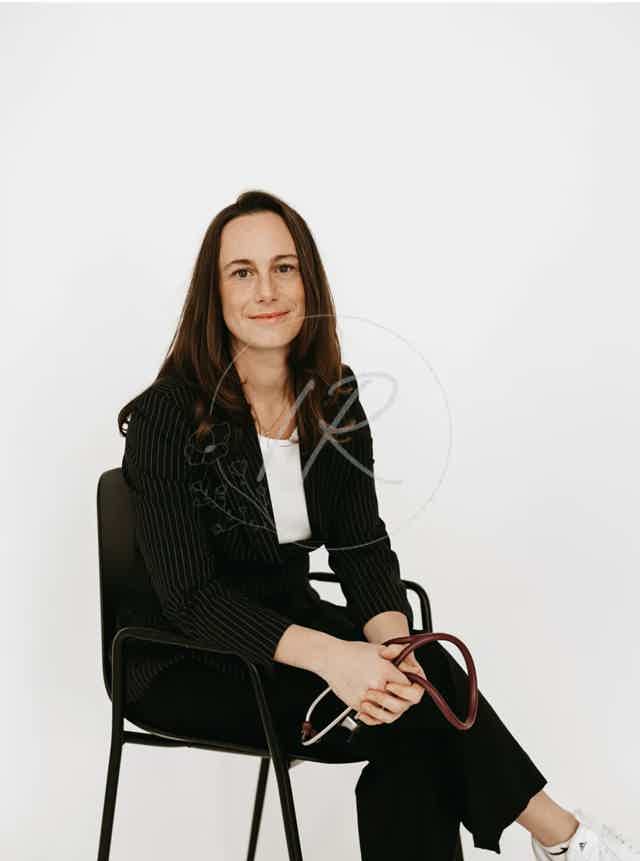 Smiling woman seated in black blazer, arms crossed, holding a burgundy handbag; white studio background.