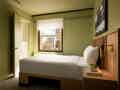 Kompaktes Hotelzimmer mit ordentlich gemachtem Einzelbett, grünen Wänden, Stadtblick und einem Nachttisch mit Pflanze und Büchern.