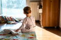 Toddler in patterned pajamas sits on a colorful bedspread, gazing out a sunlit window in a cozy room with scattered books and an open drawer.
