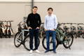 Deux hommes debout, chacun devant un vélo, dans un atelier vélo avec d'autres cycles derrières