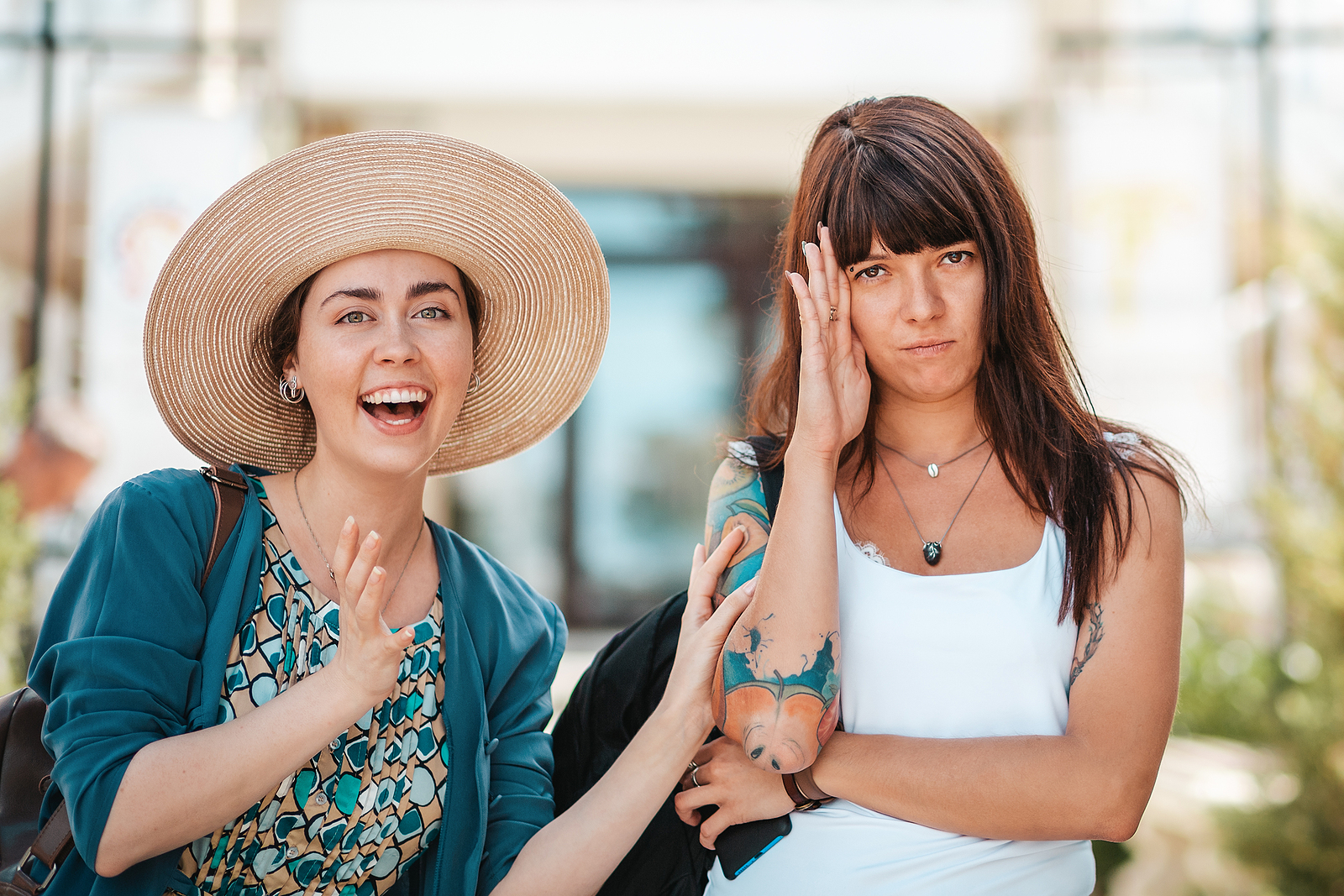 Photo of two women one is smiling holding onto her friend while the woman on the right has her hand up pretending to not know her friend and annoyed.