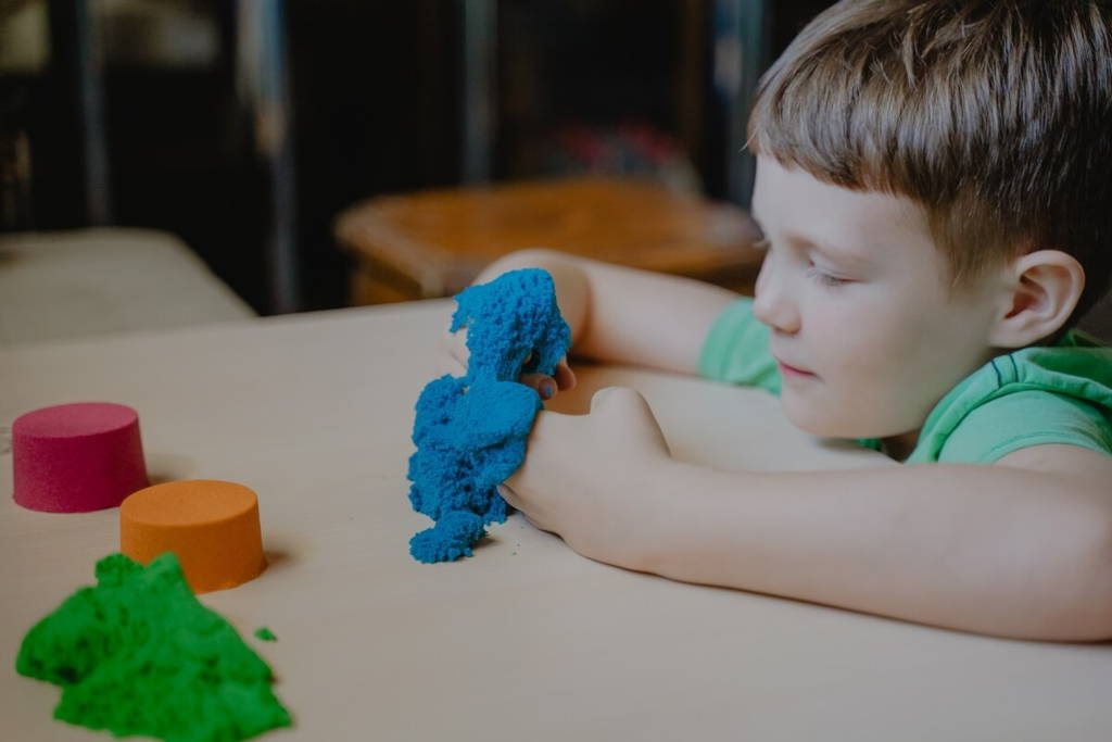 Un jeune garçon jouant avec du sable cinétique coloré à une table, pressant un paquet bleu avec ses mains tandis que d'autres couleurs - vert, orange et rose - sont à proximité.