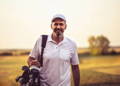 Smiling senior man carrying golf bag with clubs on shoulders at outdoor golf course.