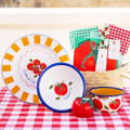 Talking Table bright tomato collection of picnic ware, displayed on a red check table cloth. Tomato illustrations against a white background. A large plate with mustard coloured stripes around rim and a bunch of cherry tomatoes in the centre. A bowl with a blue rim and a single salad tomato illustration in centre. A small bowl with multiple salad tomato illustrations around outside, a blue rim and mustard yellow colour interior. A candle in a large tomato and a straw basket containing a tomato napkin pack and green and red check cotton napkins.