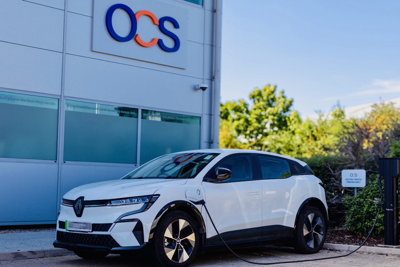 A white electric car is charging at a charging station outside a modern building with the OCS logo on the wall. Trees and greenery are visible in the background on a sunny day.