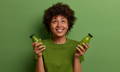 African-American girl in a green shirt, holding a glass bottle of green smoothie in each hand, in front of a green background