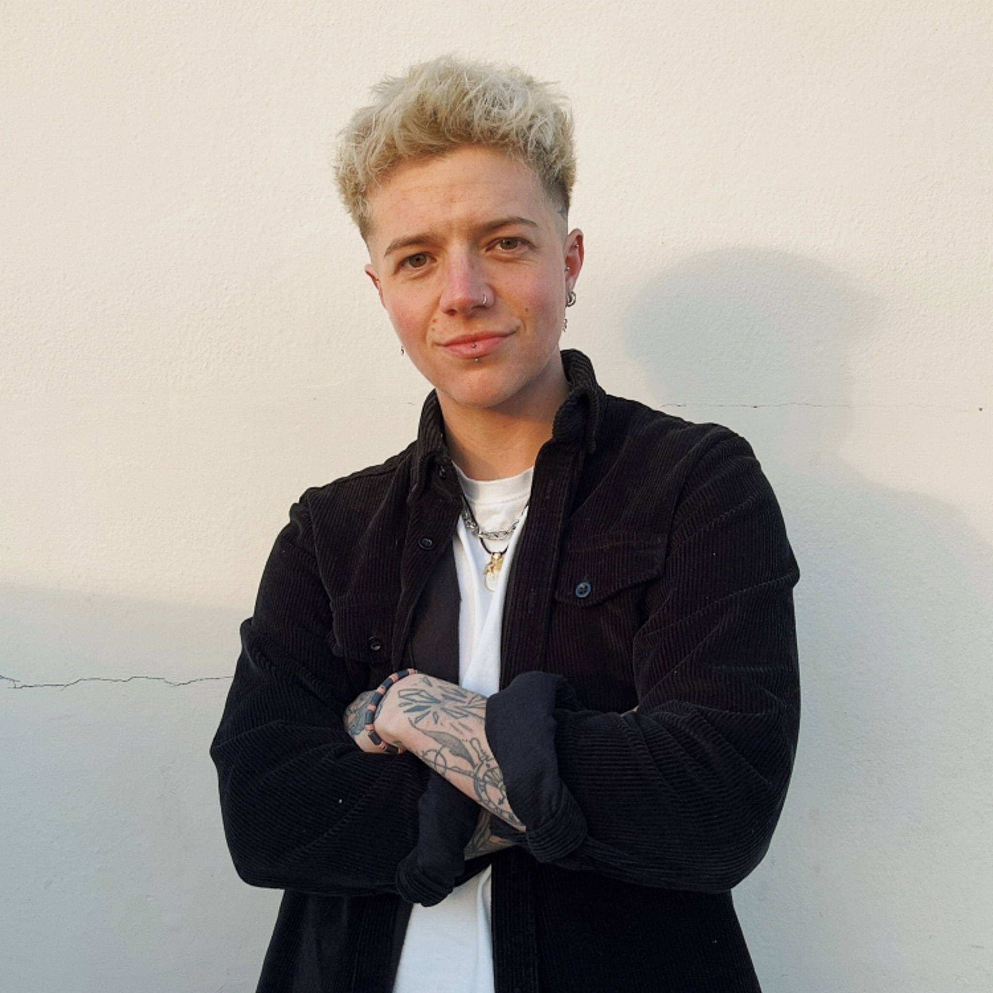 Model TJ Lucas stands against a white background with their arms folded wearing a black jacket over a white t-shirt. Arm tattoos can be seen from the rolled-up sleeve.