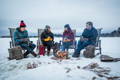 4 people sitting around a fire in the snow talking with packs resting next to each chair