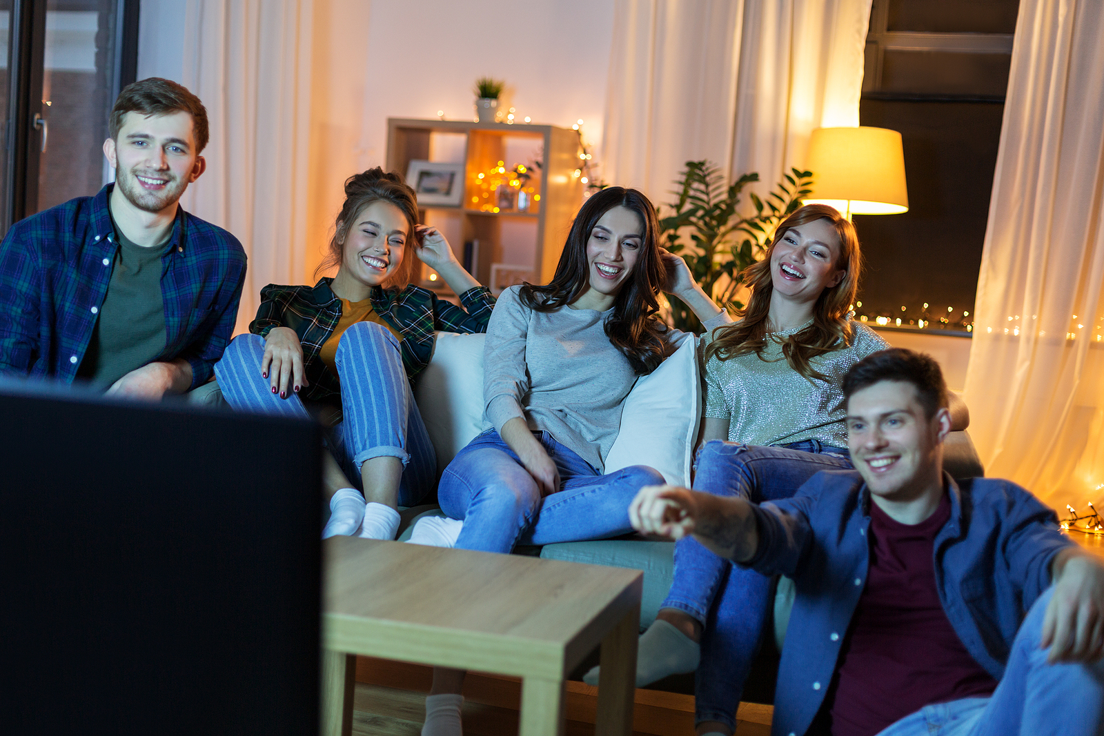 Group of 5 friends watching tv together sitting on the couch laughing.