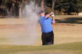 A senior male golfer wearing a blue shirt and glove swings an iron to splash a ball out of a sand bunker.