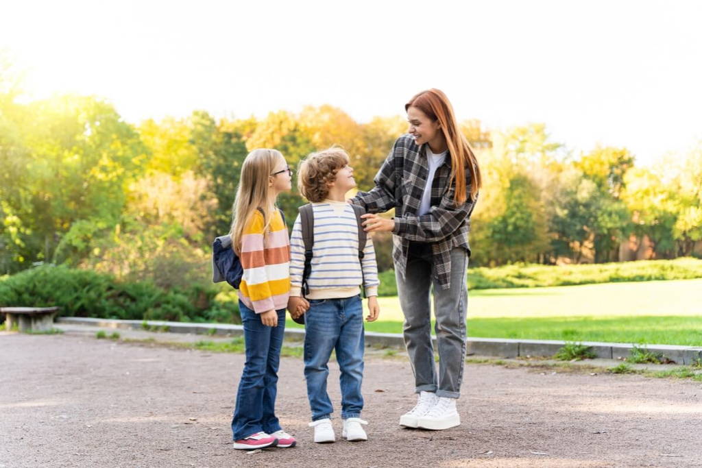 A young woman talking with two smiling school children outdoors, with all three standing on a path in a park surrounded by greenery and warm sunlight.